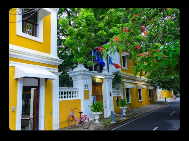 A vibrant yellow heritage building on a calm Pondicherry street with lush green trees and a parked bicycle
